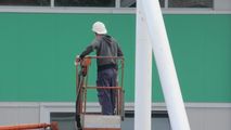Worker in a white hard hat standing on a lift beside a green building wall.
