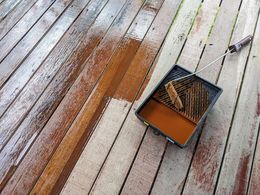 Paint roller tray with brown paint on a weathered wooden deck, surrounded by paint splatters.