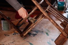 A person brushes varnish onto a wooden chair frame on a worktable.
