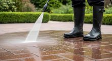 Person in black boots pressure-washing a wet brick walkway outdoors