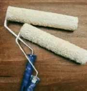 Two paint rollers with white paint on a wooden surface, one with a blue handle