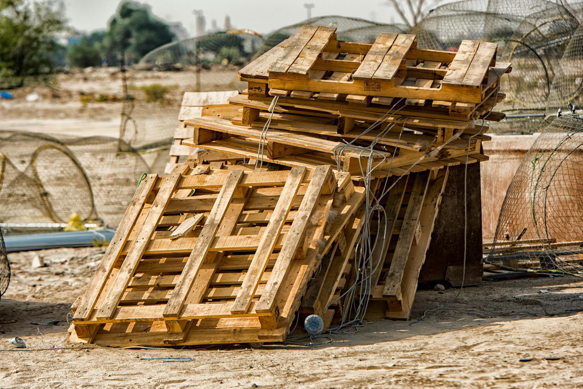 Stacks of wooden pallets outdoors, next to a blue dumpster and metal container, under a cloudy sky.