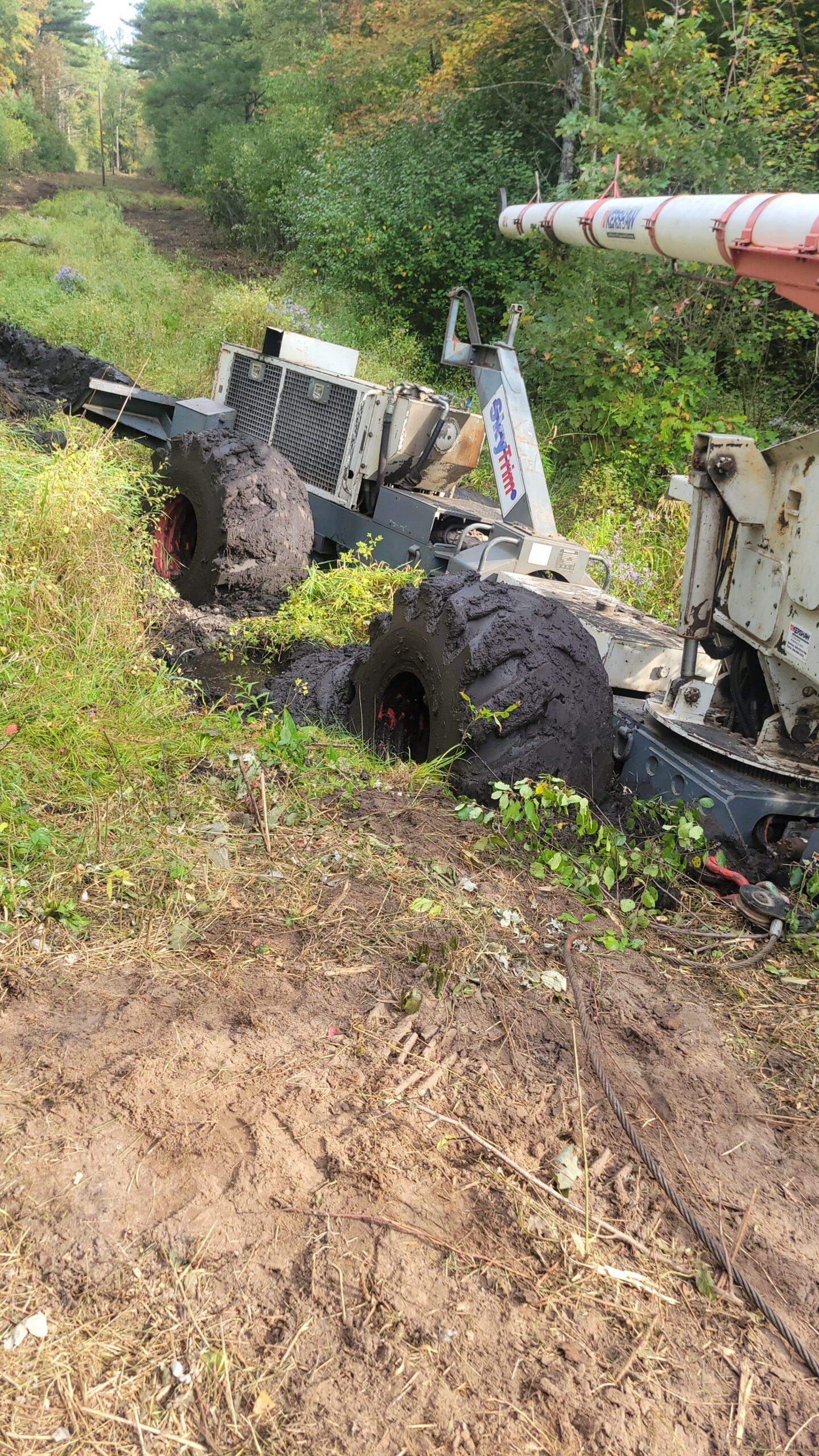 Overturned utility truck in muddy ditch beside a green roadside slope, with a tilted metal pipe attached.