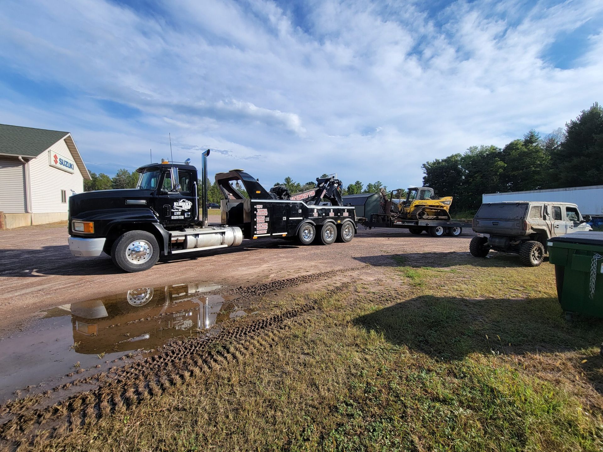 Black tow truck hauling a car carrier on a muddy roadside with puddles and a house nearby