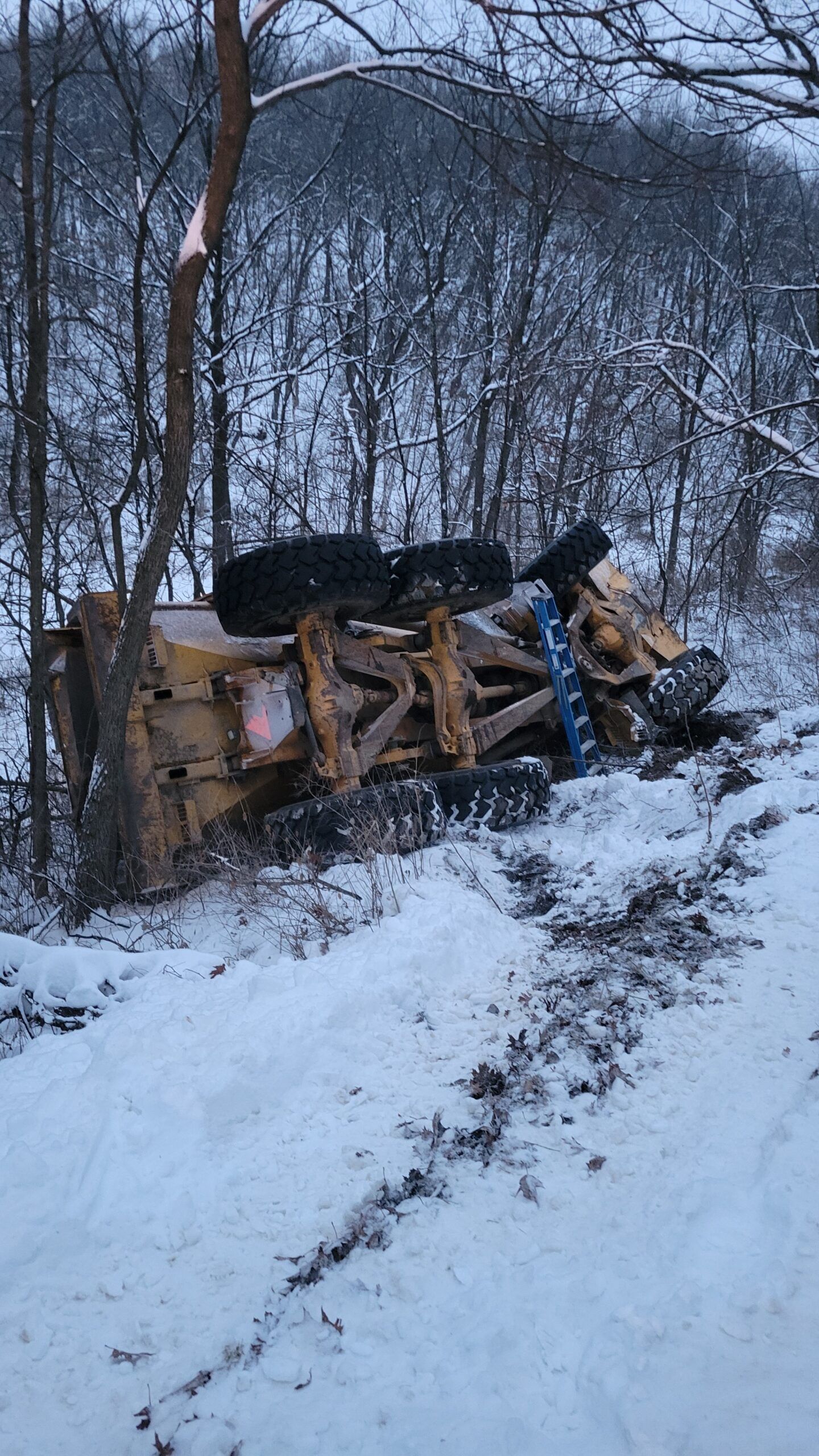 Overturned yellow vehicle in snowy woods beside a muddy track