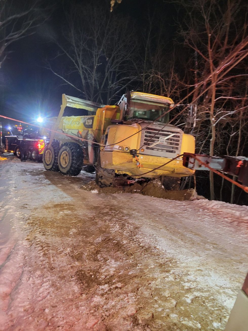 Yellow dump truck stuck in snow on a dark roadside, with emergency lights nearby