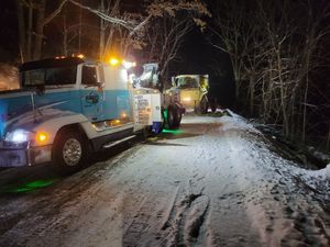 Snowy road at night with a blue utility truck and work lights clearing the path