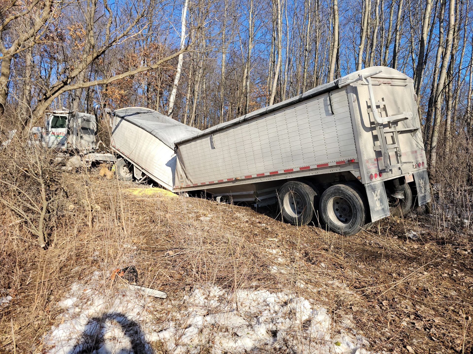 Overturned white semi-trailer stuck on a wooded hillside with bare trees and muddy ground