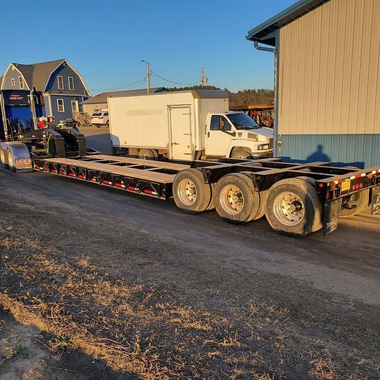 Flatbed trailer hauling a white box truck on a gravel road beside buildings at sunset