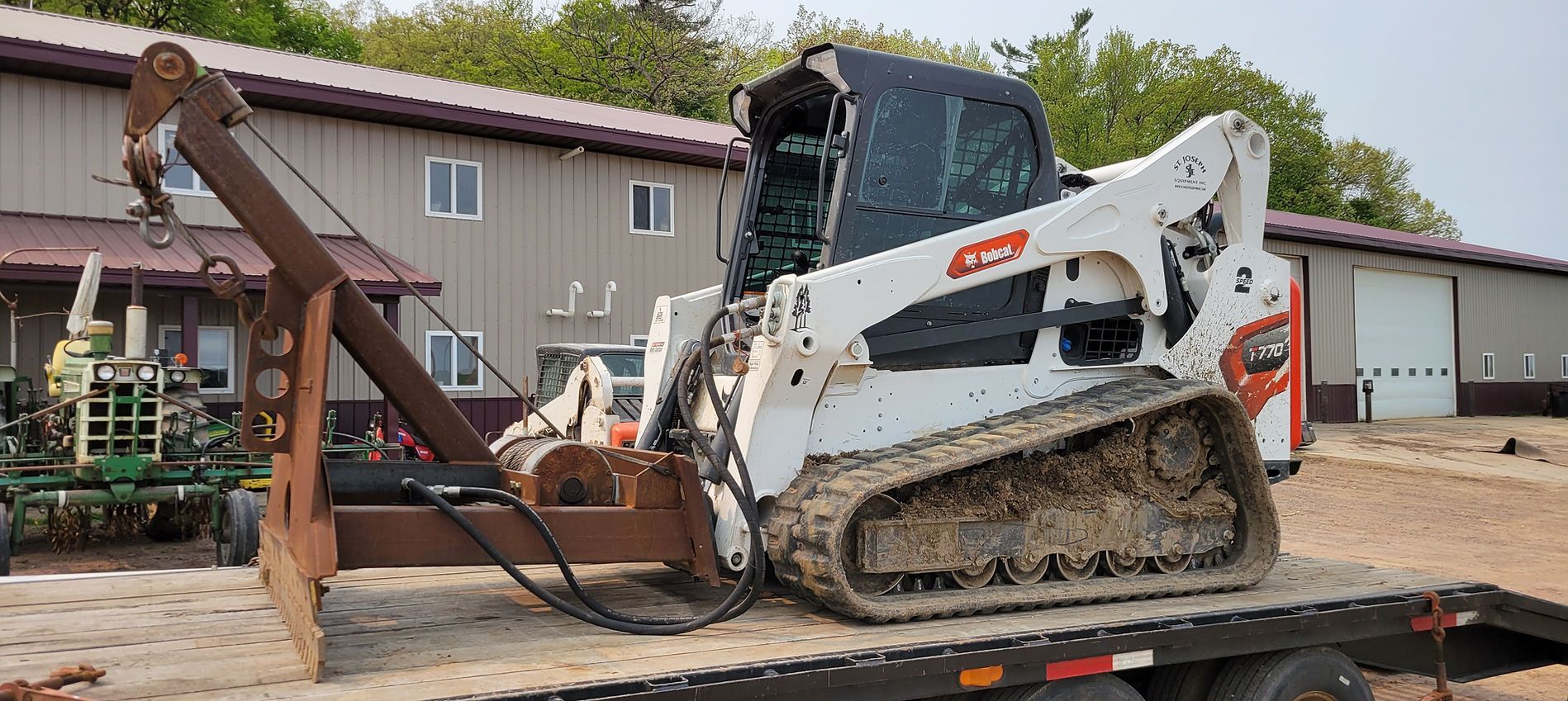 White skid-steer loader on a flatbed trailer beside a brown tow truck in an industrial lot