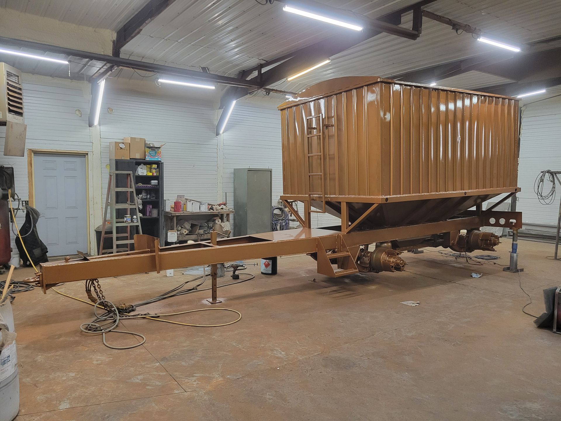 Wooden trailer frame with a small cabin in a workshop, viewed from the side.