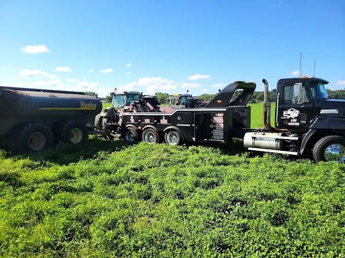 Two black and green trucks with a tanker and flatbed parked in a grassy field under a blue sky.