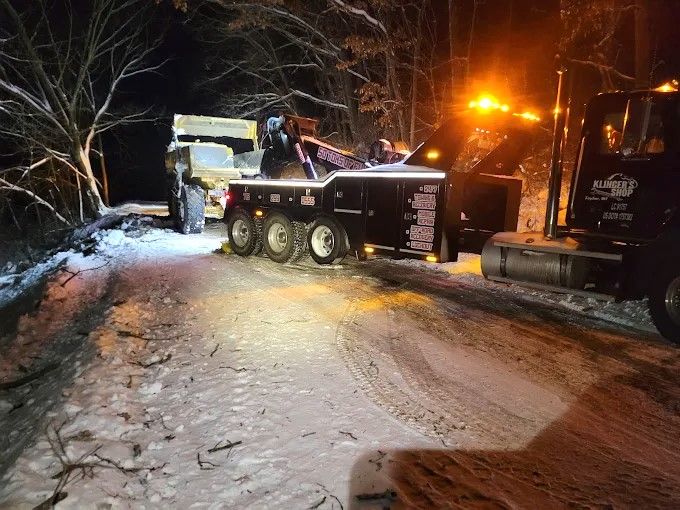 Tow truck and utility truck on snowy road at night, with orange lights illuminating the scene.