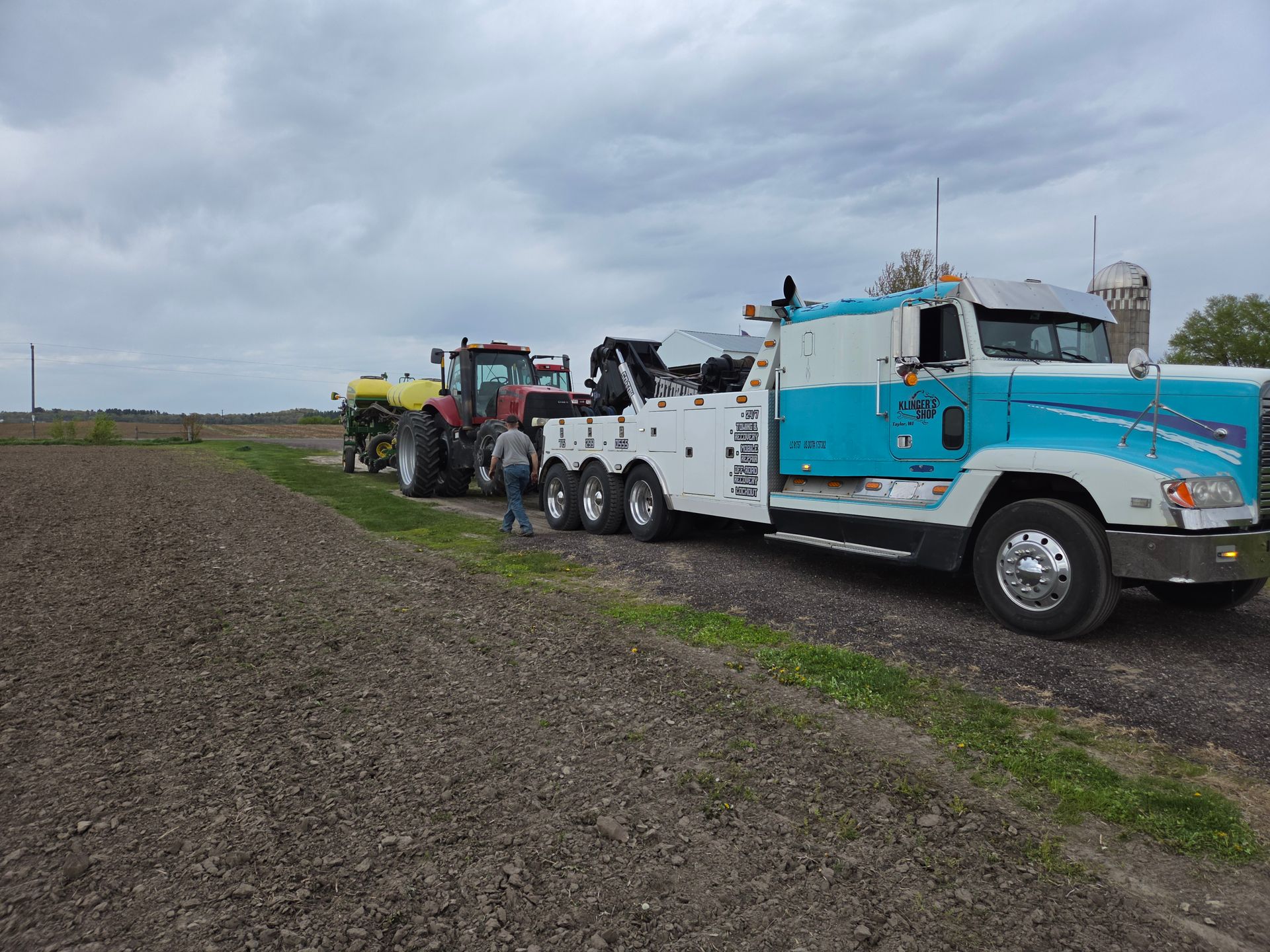 Blue tow truck and tractor parked beside a field under a cloudy sky