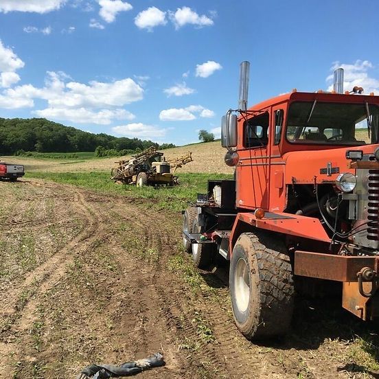 Orange farm truck beside a field with a farm machine working in the distance under a blue sky