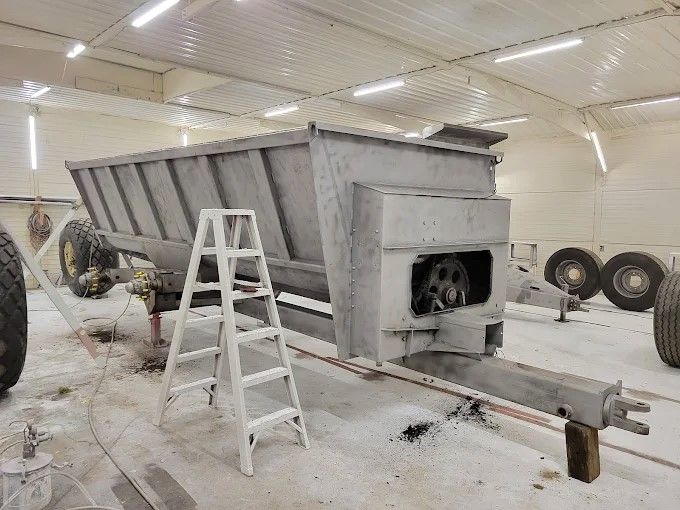 Large gray dump trailer in a workshop, with a white ladder beside it and spare wheels in the background
