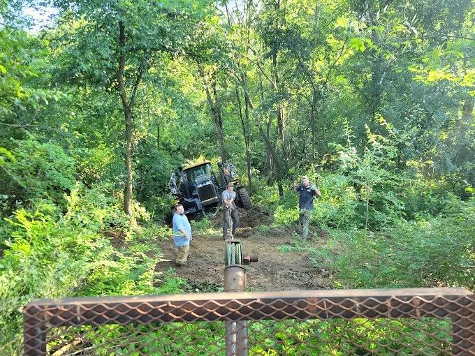 Three people near a small off-road vehicle on a forest trail, with a metal bridge railing in the foreground.