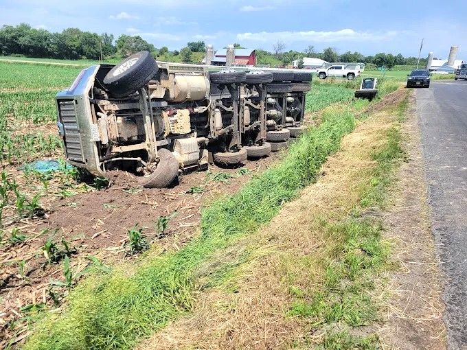 Overturned truck on its side beside a rural road, with debris and another vehicle in the distance.