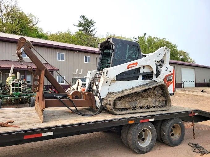 White skid steer with grapple bucket on a flatbed trailer in a yard beside a workshop
