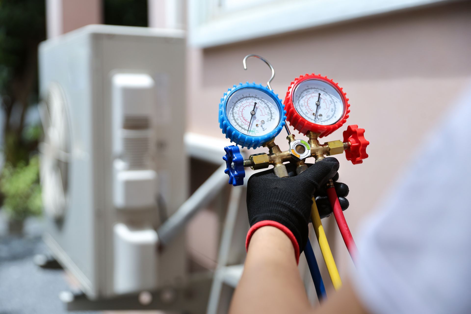 Gloved hand holding blue and red pressure gauges beside an air-conditioning unit