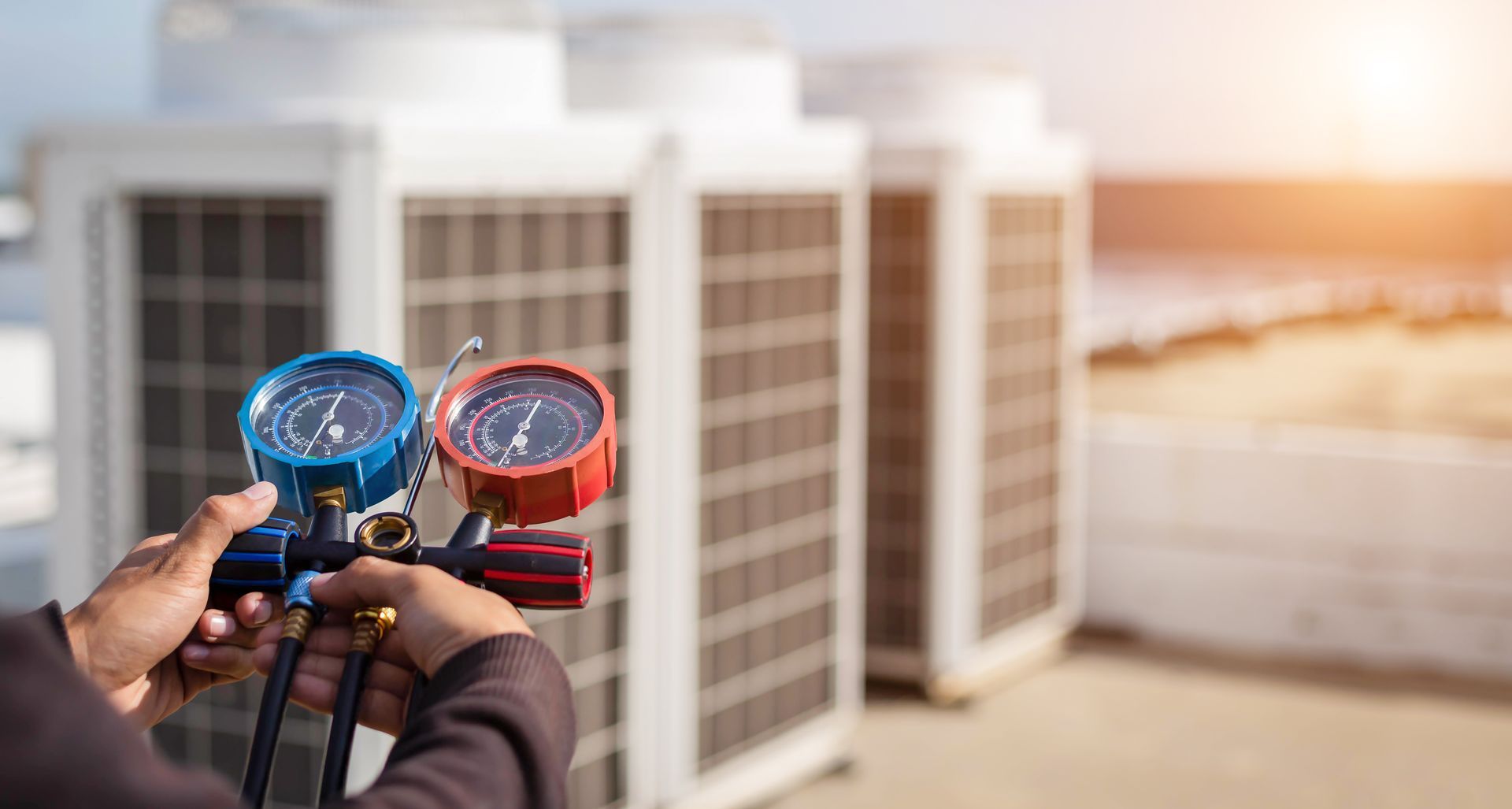 Hands holding two colored pressure gauges beside rooftop HVAC units at sunset