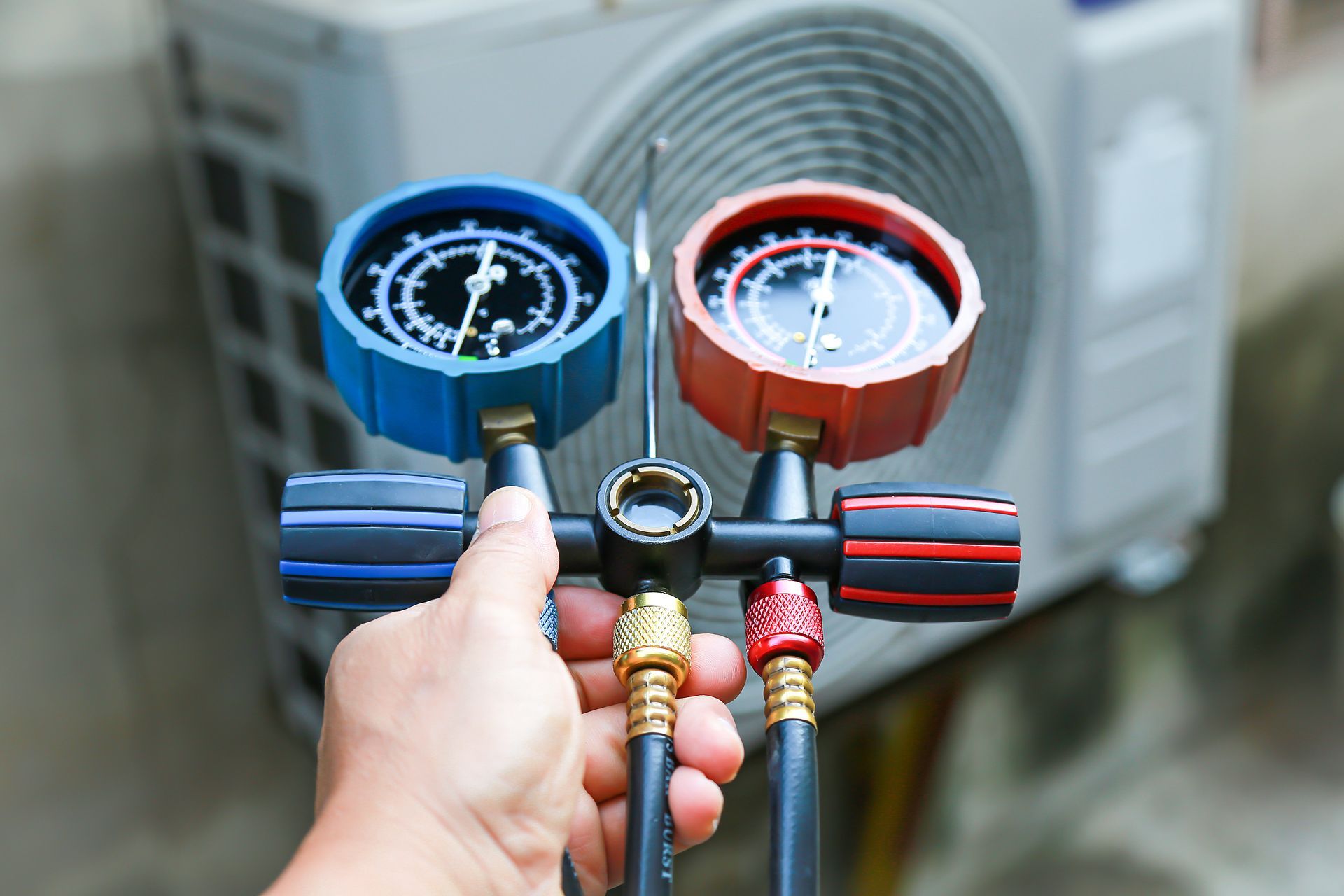 Hand holding HVAC manifold gauges in front of an outdoor air conditioning unit