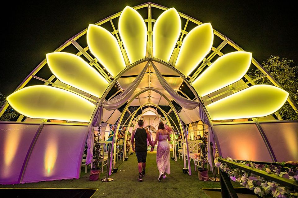 Illuminated flower-shaped entrance to the Chapel of Nature at EDC at night, with two people walking beneath it on a grassy path