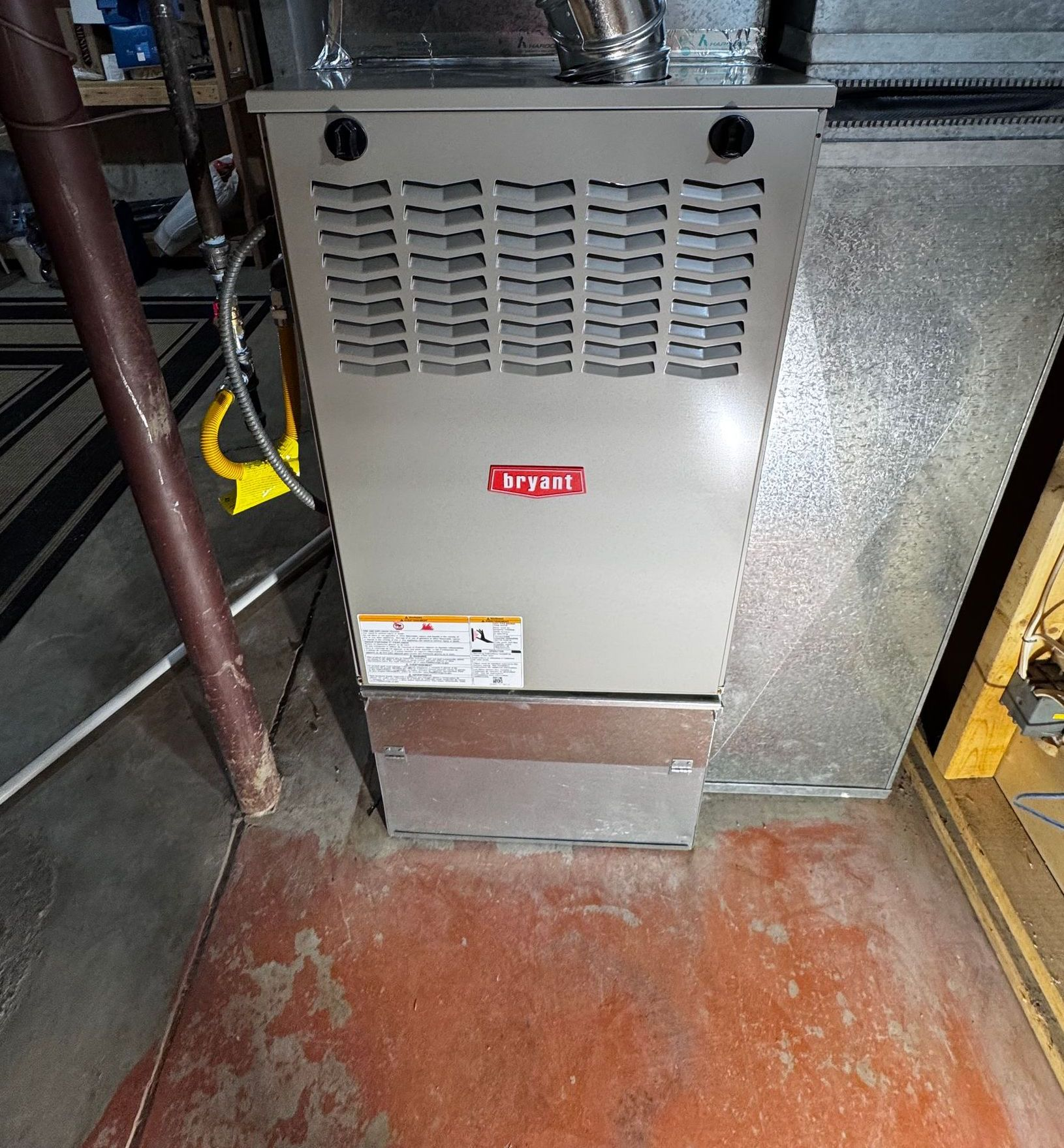 Bryant furnace in a basement setting. Gray metal unit with a front panel, ductwork visible.