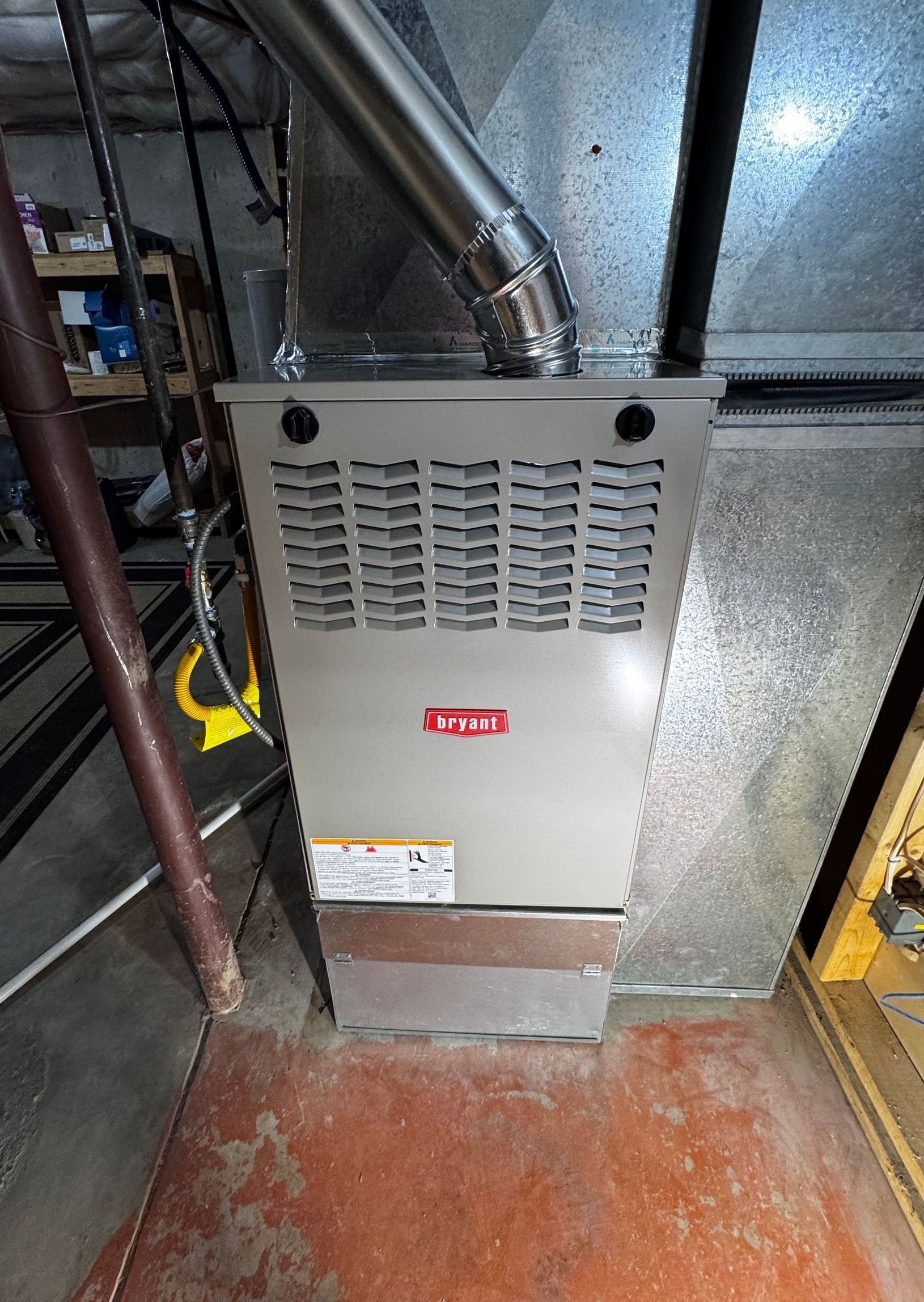 Furnace in a basement, gray metal with vent pipe, mounted on a reddish floor.