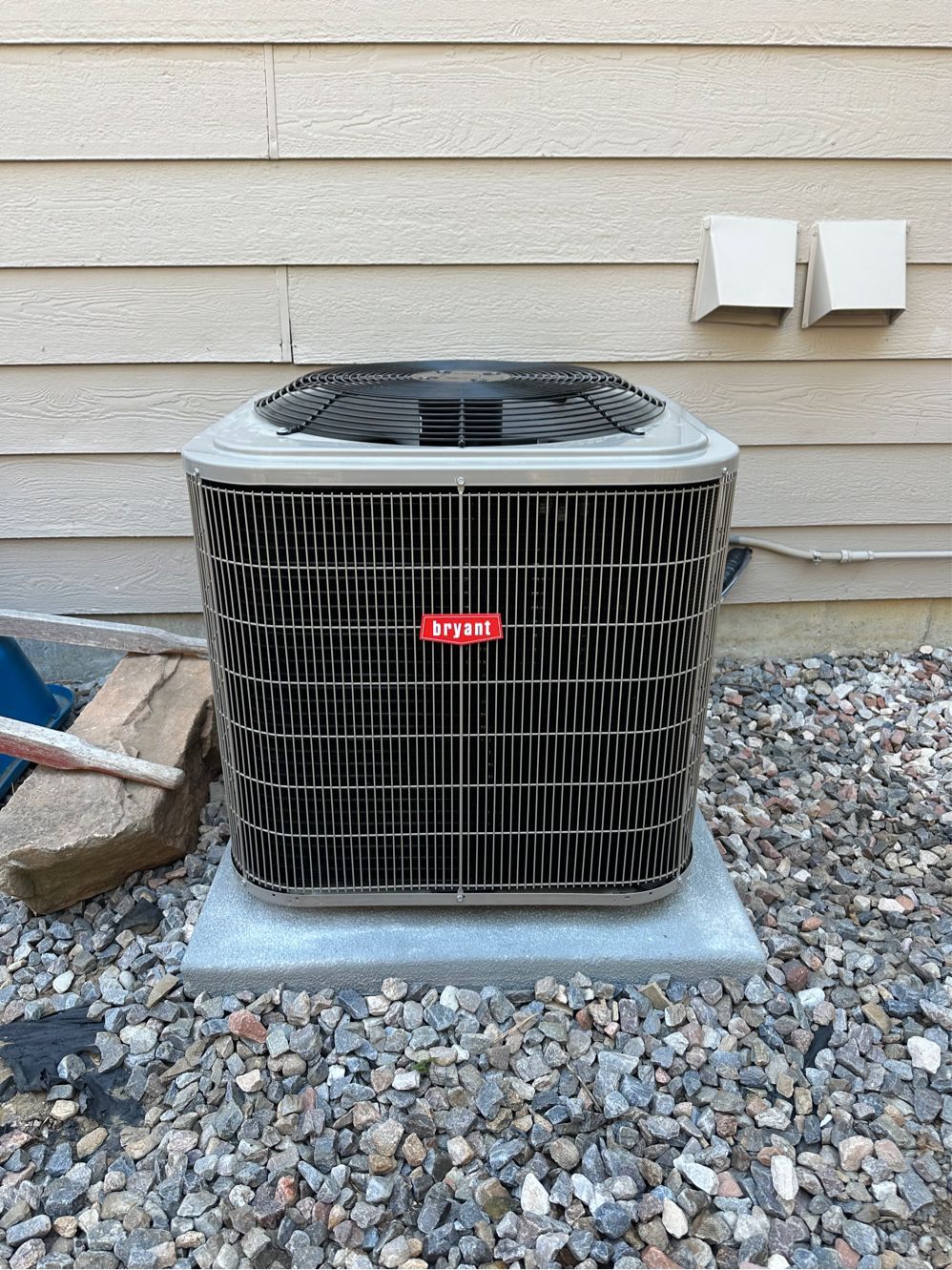 Bryant air conditioning unit on a concrete pad, surrounded by gravel, next to a beige house wall.