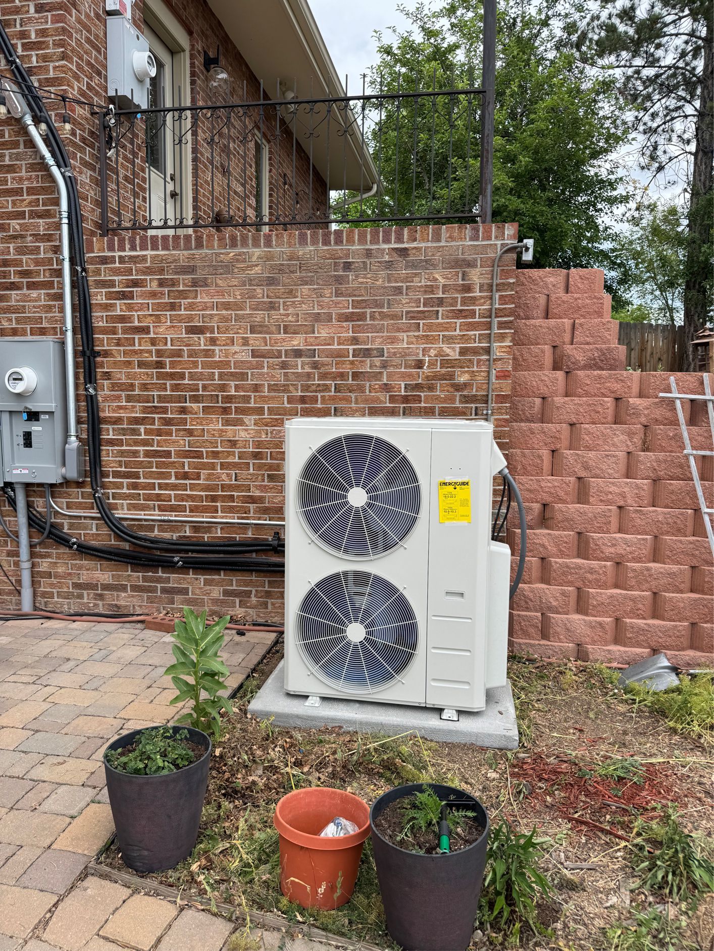 Air conditioning unit against a red brick wall, with landscaping and a retaining wall in the background.