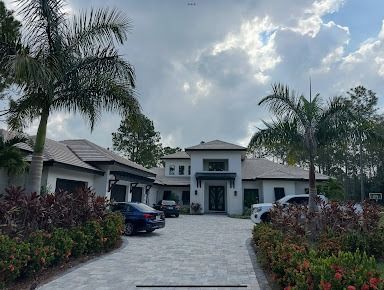 A large, modern white house with a paved driveway and palm trees, two cars parked in front.
