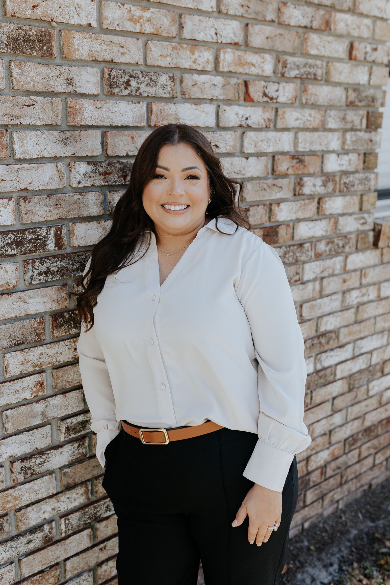 Smiling woman in a white blouse and black pants standing against a brick wall