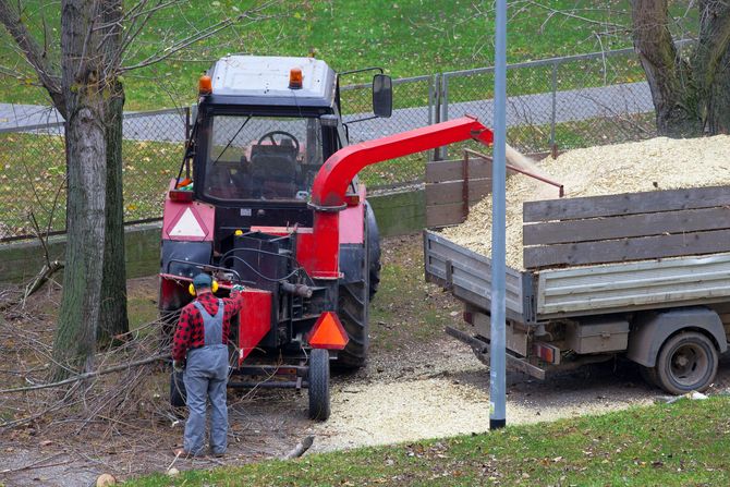 Tractor with red wood chipper beside a trailer; worker stands nearby in a wooded yard.