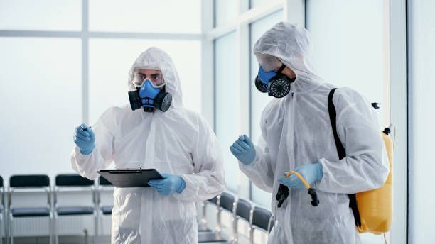 Two workers in white hazmat suits and respirators inspect a tablet in a bright corridor