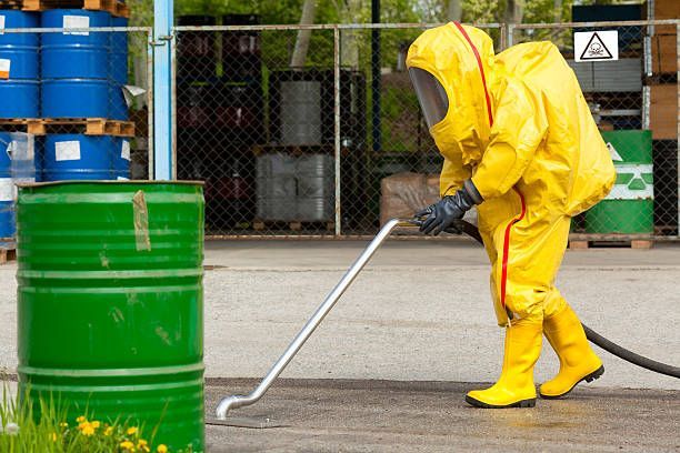 Person in a yellow hazmat suit cleaning near green and blue barrels in an industrial yard