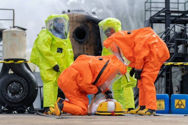 Workers in orange and yellow hazmat suits handle equipment at an industrial site near a large pipe.