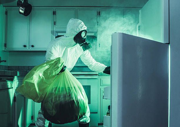 Person in protective suit cleaning a kitchen with a spray bottle beside an open refrigerator.