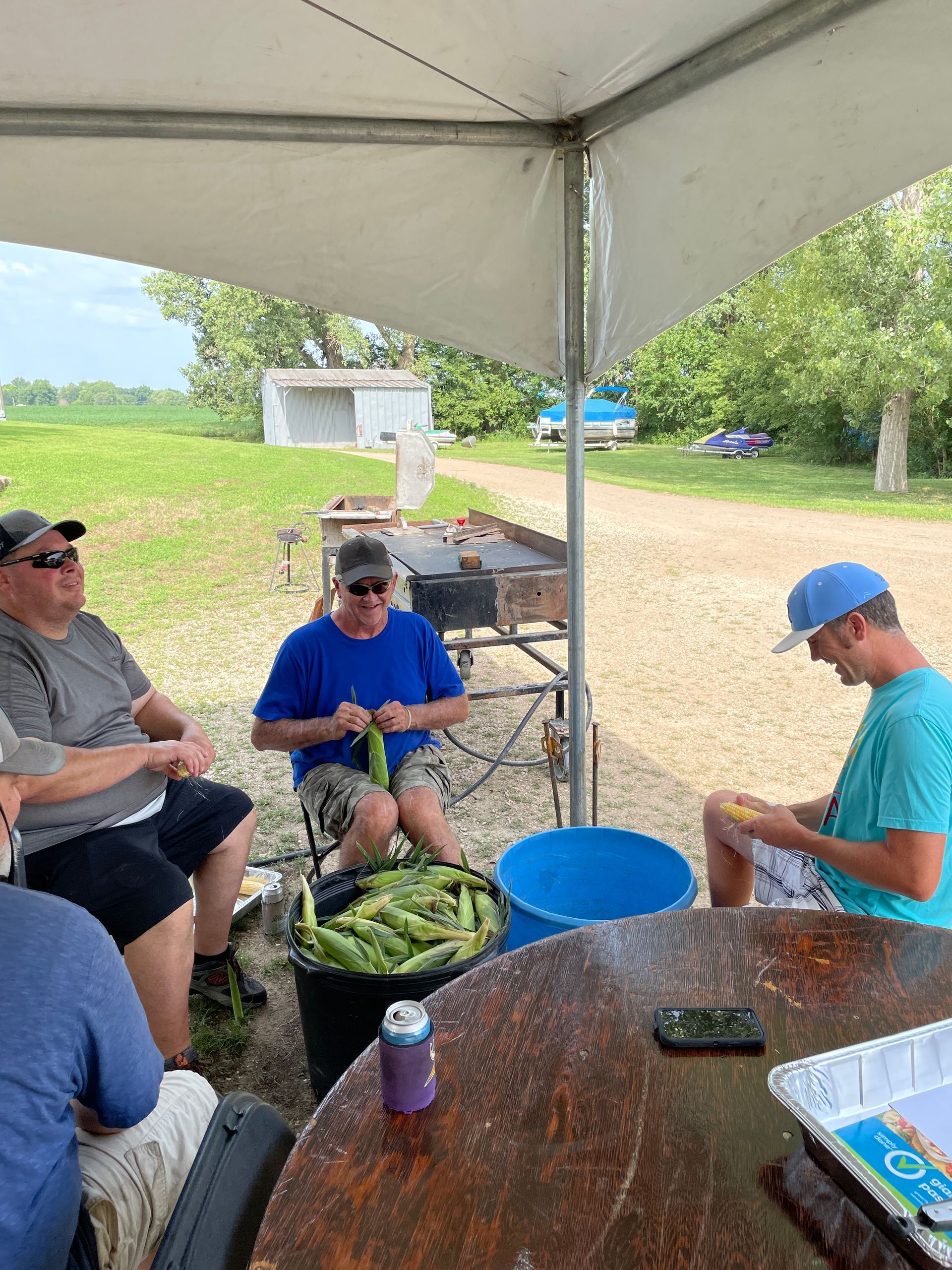 Men are sitting around a blue bucket and shucking corn at the ICL family picnic.