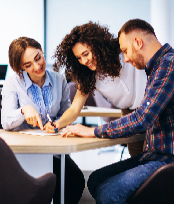 Three coworkers collaborating at a table, one pointing at a document, smiling, office setting.