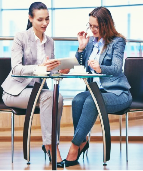 Two women in suits reviewing a tablet at a table with coffee cups.