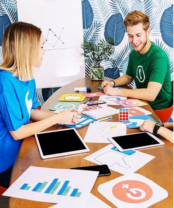 Two people collaborating at a table, looking at papers and tablets. One wears blue, the other green.