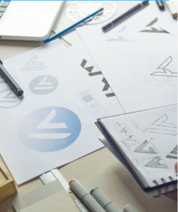 Logo designs on paper and in a sketchbook, with laptop, pens, and markers on a desk.