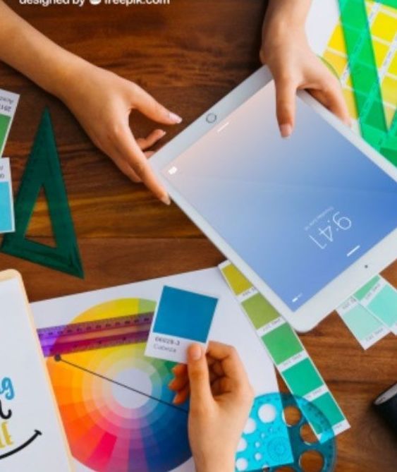 Hands holding an iPad and color swatches on a wooden table, likely a designer's workspace.