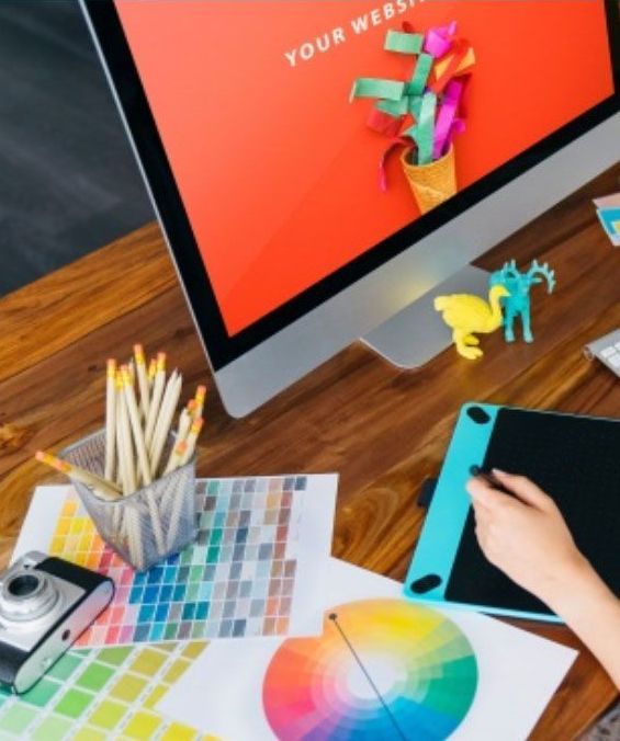 Desk with computer, design tools, color palettes, camera. A hand uses a stylus on a tablet.