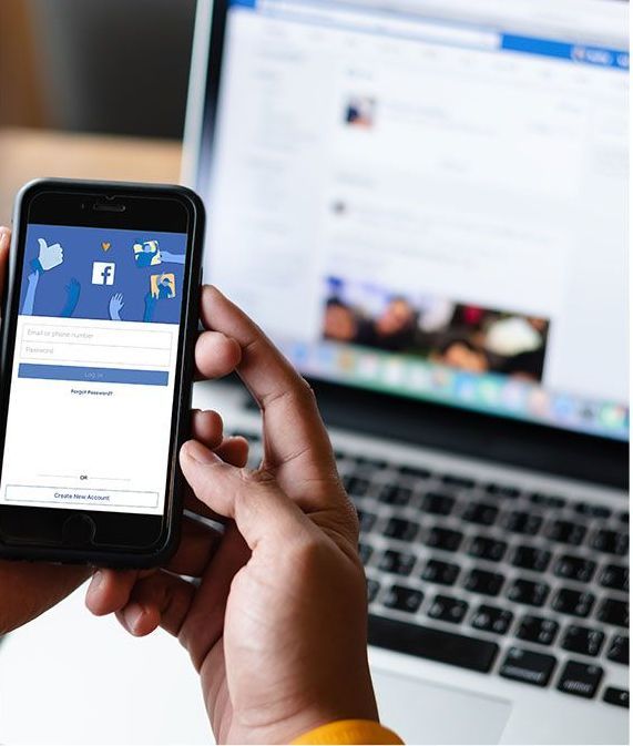 Person holding a phone displaying a Facebook login screen in front of a laptop with Facebook open.