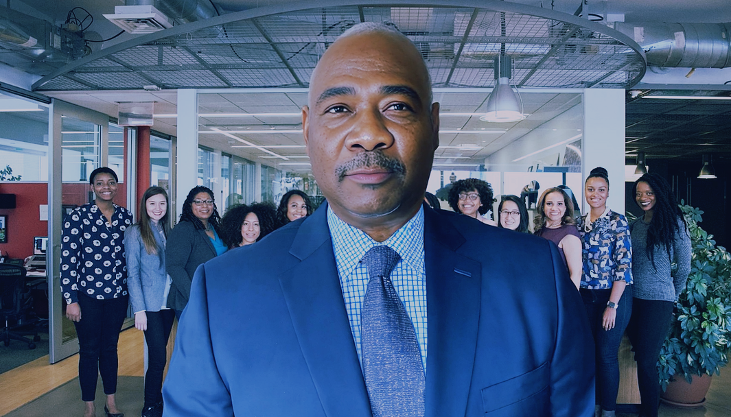 Man in suit, with diverse group of women behind him in an office setting.