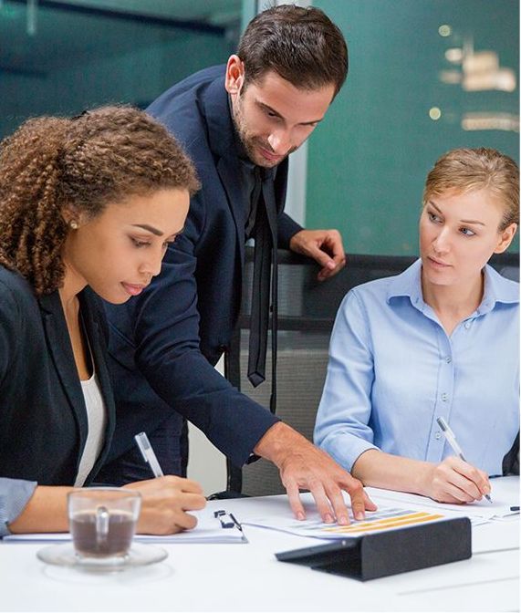 Three businesspeople review documents at a table. The man points, while two women write.