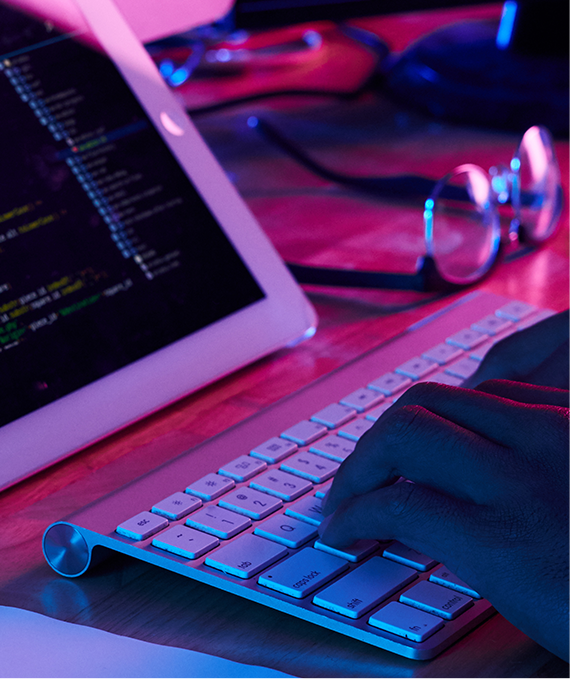 Hands typing on a keyboard with a tablet displaying code in a dark environment lit with purple and pink hues.