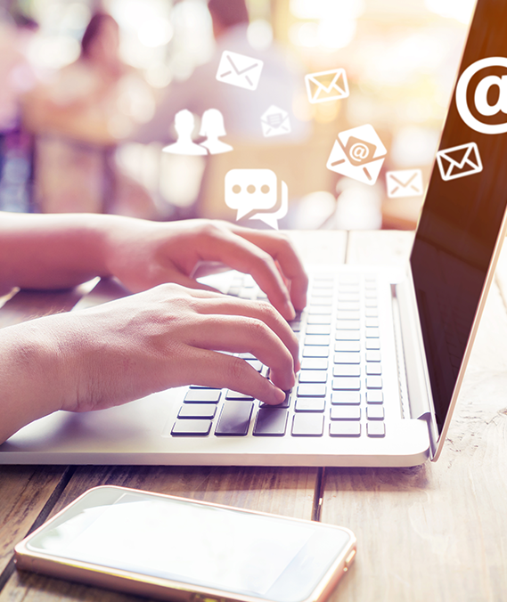Person typing on laptop with email icons above, phone on table.